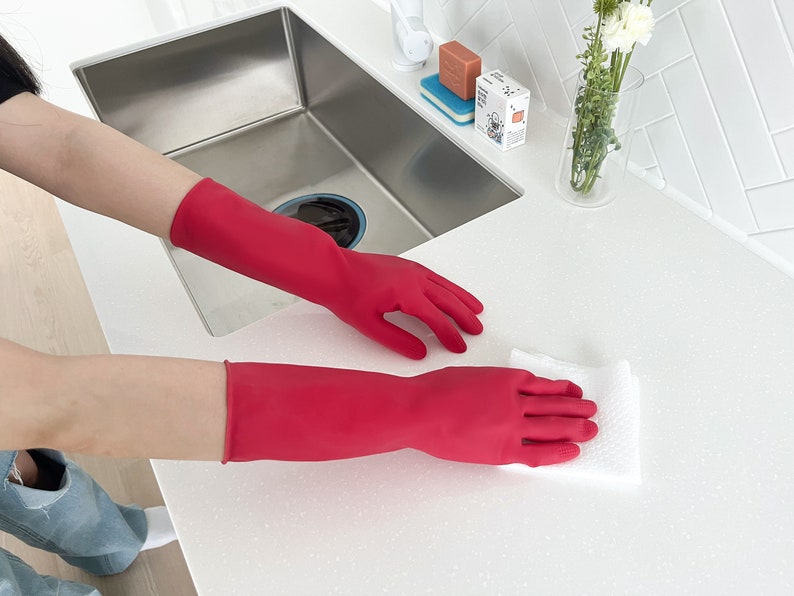 May include: A person wearing red rubber gloves is cleaning a white countertop with a white cloth. The countertop is next to a stainless steel kitchen sink.