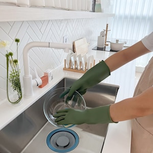 May include: A person wearing green rubber gloves washes a glass bowl in a stainless steel kitchen sink. The sink has a blue drain stopper.