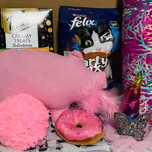 May include: A collection of cat toys and treats, including a pink pig-shaped plush, a donut toy, a fluffy ball, and a Purina Felix cat food bag. A decorative unicorn and a butterfly are also included.