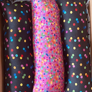 Puede incluir: Tres juguetes para gatos en una caja de cartón. Dos son negros con lunares coloridos y uno es rosa con un patrón de confeti. Los juguetes son rectangulares y parecen estar rellenos de hierba gatera.