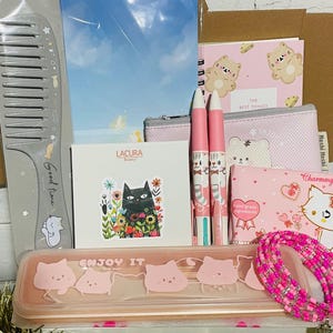 Puede incluir: Una colección de artículos de papelería y accesorios rosas y blancos, que incluyen bolígrafos, un peine, un cuaderno, un estuche y una pulsera de cuentas. Los artículos presentan diseños de gatos y osos, con las palabras "Good Time" y "Enjoy It" visibles.
