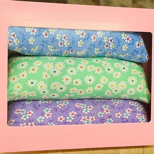 Puede incluir: Tres almohadas de tela con estampados florales. La almohada superior es azul claro con flores blancas, la almohada del medio es verde claro con flores blancas y la almohada inferior es morada con flores blancas.