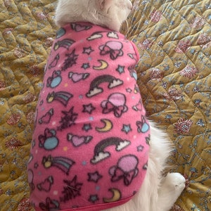 May include: A white cat wearing a pink fleece sweater with a colourful print of stars, hearts, rainbows, and clouds. The sweater has a pink trim around the neck and bottom.