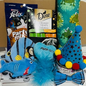 May include: A cat-themed gift box filled with treats and toys. Includes a blue fish and shark plush, a heart-shaped toy with "Purr-fect" text, a party hat, a bow tie, cat food, and cat treats.