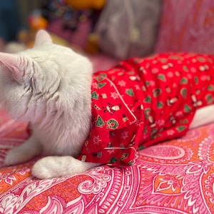 May include: A white cat wearing a red and green Christmas-themed pajama shirt, lying on a pink and orange patterned bedspread.
