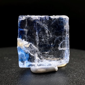 May include: A rectangular crystal with a blue tint, displayed on a small, pale stone. The crystal's surface shows cracks and flaws. The background is dark. The crystal is a mineral specimen.