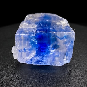 May include: A raw, crystalline mineral specimen with a striking blue and white color gradient. The stone is illuminated, showcasing its translucent qualities and natural, irregular form. The background is a dark, solid color.