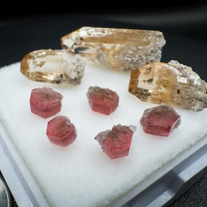 May include: A collection of three elongated, translucent amber-coloured crystals and four smaller, hexagonal, deep pink crystals arranged on a white foam display pad, with a portion of a silver-coloured coin visible in the lower left corner.