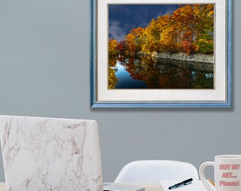 May include: A framed landscape photograph featuring a serene lake reflecting vibrant autumn foliage in shades of red, orange, and yellow. The photograph is set in a blue frame and hangs on a gray wall. A laptop, notepad, pen, and a mug with text are on a wooden desk.