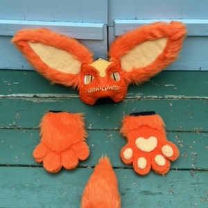 May include: An orange and yellow furry dragon mask with large ears, and matching orange furry paw gloves and tail. The mask has a detailed face with eye openings and a toothy grin. The gloves have white paw pads. The tail is cone-shaped.