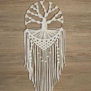 Peut inclure: Une suspension murale en macramé blanche avec un cercle en métal doré et des perles en bois. Le macramé est en forme d'arbre avec une canopée circulaire et de longues mèches qui pendent.