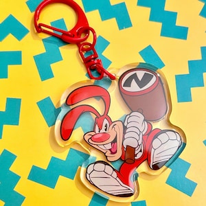 May include: A clear acrylic keychain featuring a cartoon rabbit character in red and white, holding a brown mallet. The keychain has a red clasp and ring. The background is yellow with blue geometric shapes.