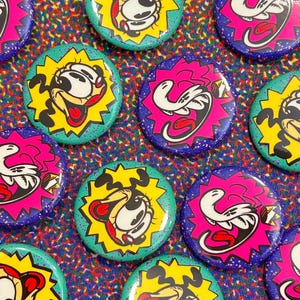 Puede incluir: Una colección de botones redondos y coloridos con diseños de personajes de dibujos animados. Los botones presentan un personaje parecido a un perro con un fondo de explosión amarillo y un personaje blanco con un fondo de explosión rosa. Los botones están dispersos sobre una superficie estampada.