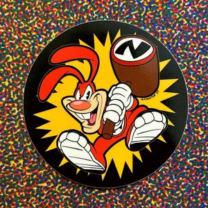 May include: A round sticker featuring a cartoon rabbit in a red jumpsuit, white gloves and shoes, and a brown bat. The rabbit is smiling and holding the bat, with a brown barrel and the letter "N" in the background. The sticker has a yellow starburst design.