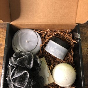 May include: A gift box filled with bath and body products. The box contains a black and white bath pouf, a white bath bomb, a gray candle, and a gray jar of bath salts.