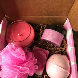 May include: A pink and white gift box filled with bath and body products. The box contains a pink candle, a pink bath bomb, a pink bath puff, a pink soap bar, and a pink jar of body scrub.