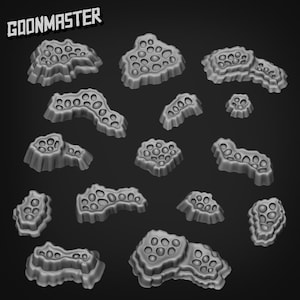 Puede incluir: Una colección de piezas de terreno grises y porosas, posiblemente para juegos de mesa o modelado. Las piezas varían en tamaño y forma, con una apariencia texturizada y orgánica. La palabra "GOONMASTER" está impresa en blanco en la parte superior.