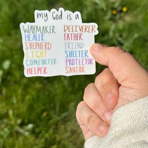 Könnte beinhalten: Ein weißer Aufkleber mit den Worten "my God is a" oben, gefolgt von Wortlisten in verschiedenen Farben. Die Wörter umfassen "Waymaker, Healer, Shepherd, Light, Comforter, Helper" links und "Deliverer, Father, Friend, Shelter, Protector, Savior" rechts.