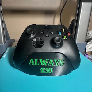 May include: Black Xbox controller with green lettering that reads "ALWAYS 420". The controller has colored buttons and two joysticks. The controller is sitting on a blue surface.