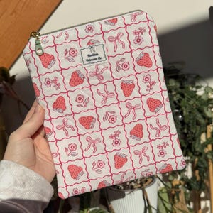 May include: A white zipped pouch featuring a pattern of pink strawberries, bows, and flowers within a red grid. The pouch has a gold zip and a small tag that reads "Bookish Princess Co."