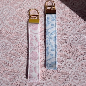 Puede incluir: Dos llaveros de tela con herrajes dorados. Uno presenta un estampado floral rosa sobre fondo blanco, y el otro un estampado floral azul sobre blanco. Los llaveros se muestran sobre una superficie de encaje rosa.