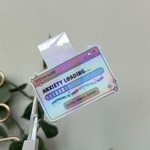 May include: A holographic sticker featuring a computer window design with the text "ANXIETY LOADING...". The sticker also includes the words "SYSTEM ALERT", "PREPARING TO UNZIP", and "CANCEL".