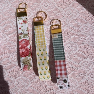 Puede incluir: Tres llaveros de tela con herrajes dorados. Los llaveros presentan estampados florales, de cuadros vichy y a cuadros en rojo, amarillo y verde. Los llaveros son de tela y tienen forma rectangular.