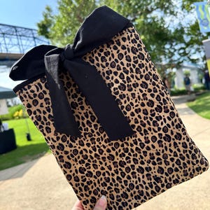 May include: A rectangular clutch bag with a leopard print pattern in brown and black. It has a large black bow at the top. The bag is held in a hand.