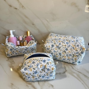 May include: Three floral print cosmetic bags and a small basket. The bags and basket are white with blue and yellow flowers. The bags are different sizes and shapes. The basket holds pink bottles and a soap dispenser.