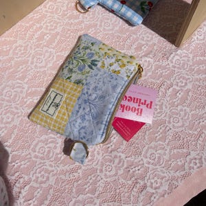 May include: A patchwork zippered pouch with floral and gingham patterns, a gold zipper, and a small ring. A pink card with the text "Prince Book" is partially visible. A matching cherry-print pouch is in the background.
