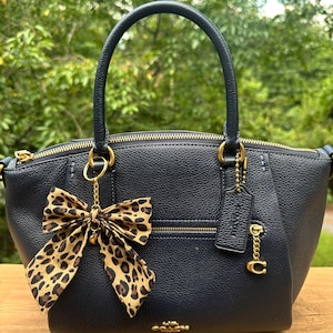 Puede incluir: Un bolso de mano de cuero azul marino con herrajes dorados, un asa superior y un lazo extraíble con estampado de leopardo. El bolso tiene cierre de cremallera y un pequeño charm con el logo de la marca.