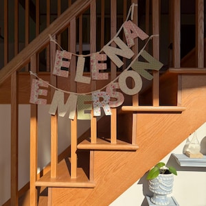 Puede incluir: Una escalera de madera con una pancarta que dice "ELENA EMERSON". Las letras de tela presentan estampados florales en rosa, verde y crema. Una pequeña planta en maceta está en un estante. La escalera es de madera.