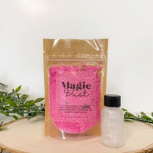 May include: A clear plastic bag filled with pink bath salts labeled "Magic Dust". The bag is sitting on a wooden surface next to a clear bottle of glitter.