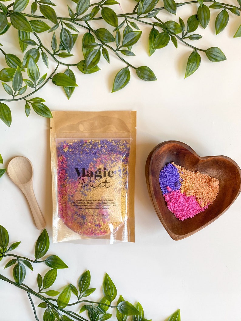 May include: A clear plastic bag filled with colorful bath salts labeled "Magic Dust". The bag is on a white surface with green leafy branches. A wooden heart-shaped bowl filled with the same bath salts is also on the surface.