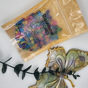 May include: A clear plastic bag filled with colorful rainbow stones. The bag is labeled "Rainbow Stones".