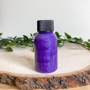 May include: A small, clear bottle filled with a shimmering, deep purple liquid. The bottle has a black cap and is set on a rustic wooden slice. Green foliage is visible in the background, adding a natural touch.