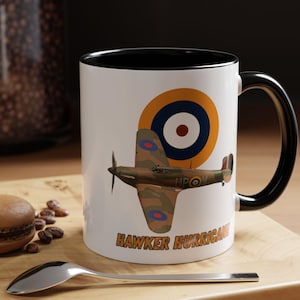 May include: A white ceramic mug with a black handle and interior rim. The mug features a graphic of a camouflaged Hawker Hurricane fighter plane in flight, positioned below a circular orange, white, and blue target emblem. The text HAWKER HURRICANE is printed in orange below the plane.