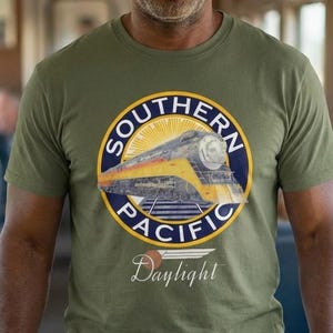 May include: An olive green t-shirt featuring a circular graphic of a yellow and blue train with the words "Southern Pacific" and "Daylight". The shirt is worn by a person standing in a train car.