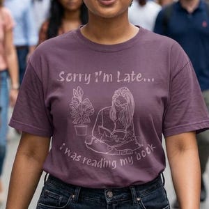 May include: A woman wearing a mauve t-shirt with the text "Sorry I'm Late... I was reading my book" and a graphic of a person reading. She is walking on a city street, surrounded by other people.