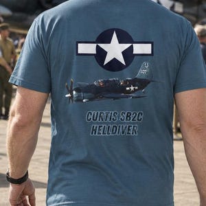 Może przedstawiać: Łupkowoniebieski t-shirt z krótkim rękawem z grafiką na plecach. Wzór zawiera białą gwiazdę w ciemnoniebieskim okręgu z poziomymi paskami, szczegółową ilustrację samolotu Curtis SB2C Helldiver oraz napis CURTIS SB2C HELLDIVER.
