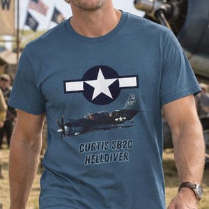 Könnte beinhalten: Ein blaues Kurzarm-T-Shirt mit einer Grafik eines Curtiss SB2C Helldiver Flugzeugs, einem weißen Stern-Emblem und dem aufgedruckten Text "CURTIS SB2C HELLDIVER" auf der Vorderseite. Das Shirt hat einen Rundhalsausschnitt und eine Standardpassform.