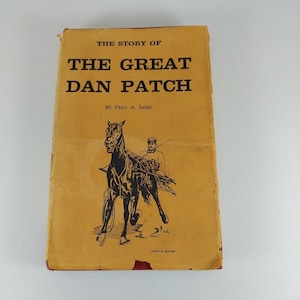 May include: A hardcover book titled The Story of The Great Dan Patch by Fred A. Sasse. The cover is mustard yellow with black text and a black ink illustration of a horse pulling a sulky with a driver. The edges show signs of wear.