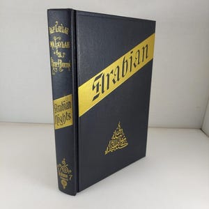 Könnte beinhalten: Ein gebundenes Buch mit dem Titel "Alf Laylah Wa Laylah" von Richard Burton, auch bekannt als "Arabian Nights". Das Buch ist schwarz mit goldener Schrift und einem goldenen diagonalen Streifen auf dem Einband. Band 7 ist auf dem Buchrücken zu sehen.