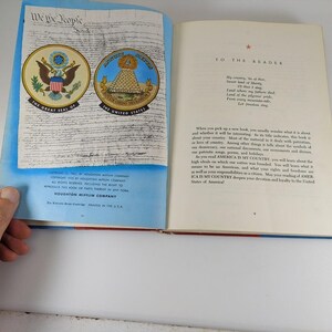 Puede incluir: Un libro abierto con una cubierta roja, blanca y azul. El libro se titula "America is My Country". Las p&aacute;ginas interiores presentan un documento escrito a mano y un sello de los Estados Unidos.