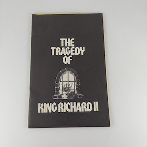 May include: A black book cover with the title "THE TRAGEDY OF KING RICHARD II" in white text. The cover features an illustration of a window with a crown inside. The book has a yellowed edge.