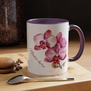 May include: White ceramic mug with a purple interior and handle, featuring a watercolor illustration of pink orchids. The mug has the text "Name here" in a cursive font. A spoon and a macaron are on a wooden surface.
