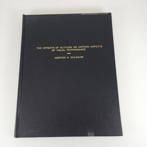 Op de afbeelding: Zwart hardback boek met gouden letters op de rug. De titel is "The Effects of Altitude on Certain Aspects of Visual Performance" door Morton K. Ohlbaum.