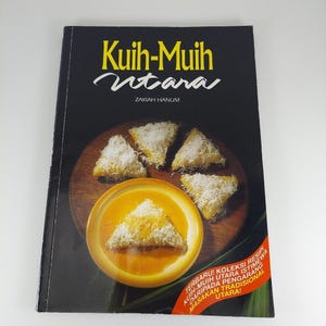 Peut inclure: Un livre de cuisine intitulé "Kuih-Muih Utara" par Zakiah Hanum. La couverture présente un fond sombre avec une assiette en bois contenant plusieurs pâtisseries triangulaires garnies de noix de coco râpée et un bol de sauce jaune.