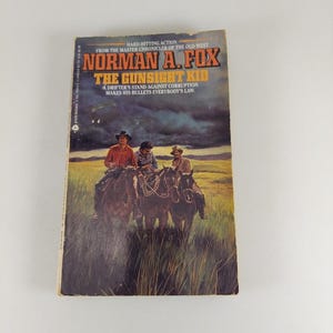 Pode incluir: Um livro de bolso intitulado The Gunsight Kid de Norman A. Fox. A capa apresenta uma ilustração de três pessoas a cavalo cavalgando por um campo de grama sob um céu escuro e tempestuoso. O texto diz Hard-hitting action from the master chronicler of the old west e A drifter's stand against corruption makes his bullets everybody's law.