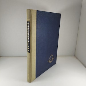May include: A hardcover book titled "Steuben Glass" with a navy blue cover and a tan spine. The spine features the title in gold lettering. A gold emblem is on the front cover. The book is in good condition.